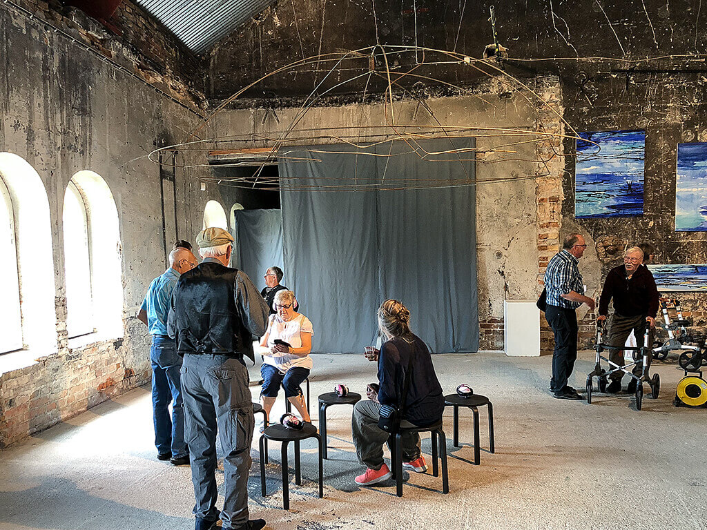 Fotografi från Ånghammaren 2018, av installation av Ulla Hägglund och Johanna Lundin som hänger från taket tillsammans med pallar under och hörselkåpor/hörlurar.